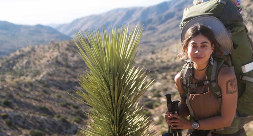 A person wearing a backpack pauses for the photo beside a desert plant. In the background, there are mountains. 